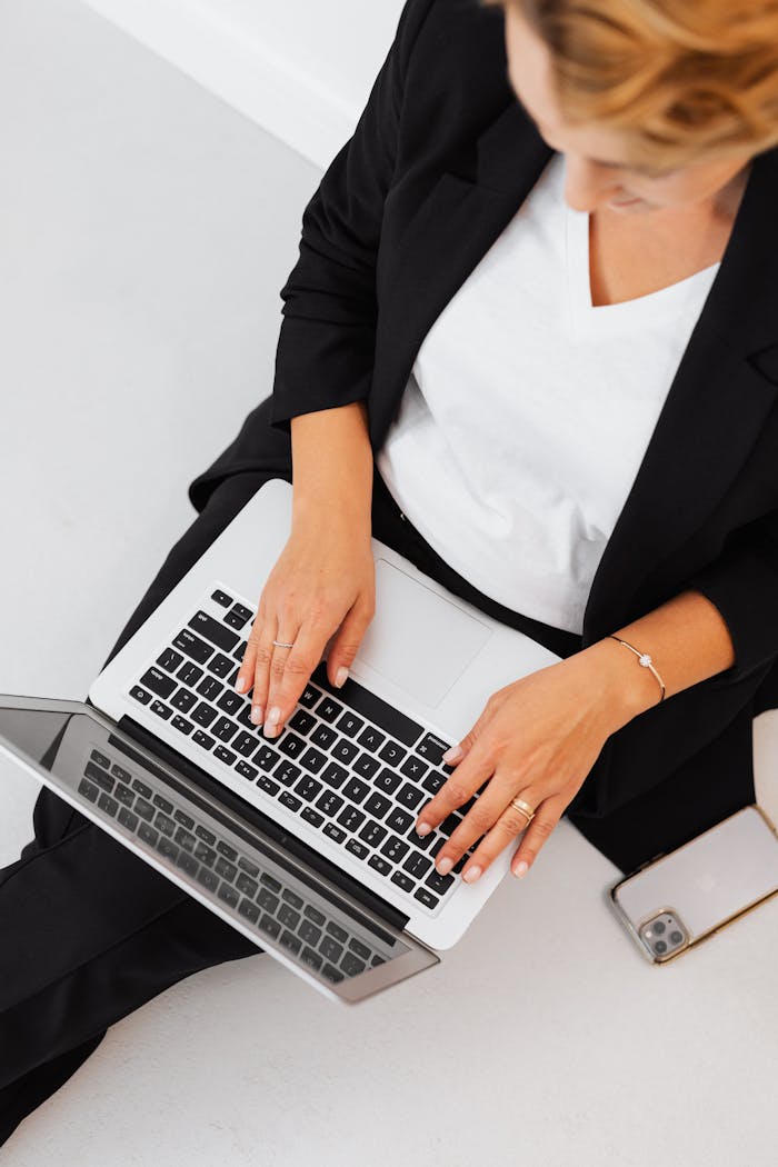 Overhead shot of a woman in a black blazer typing on a laptop with a smartphone beside her, illustrating modern technology and remote work.