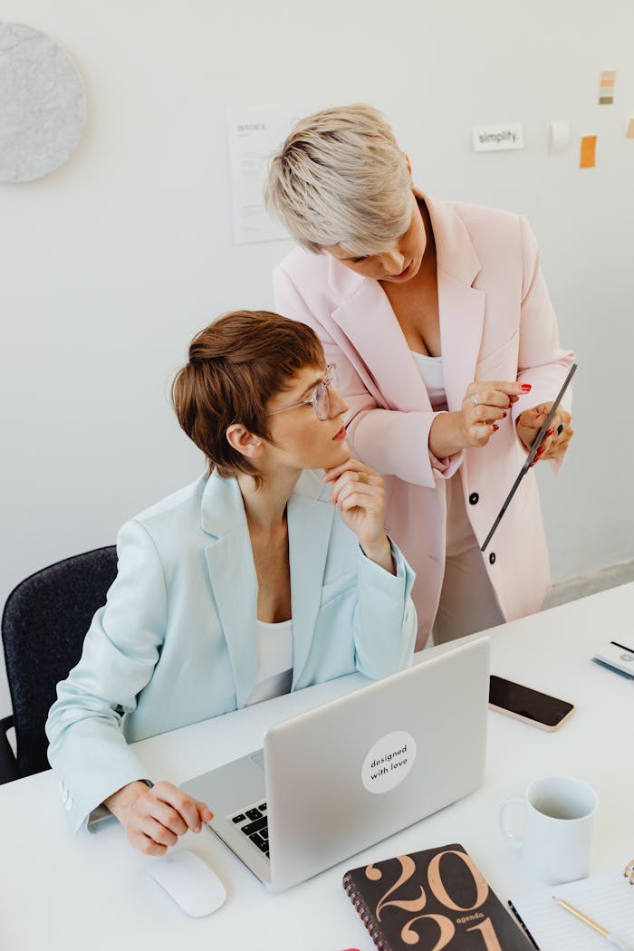 Two businesswomen collaborating over a tablet in a modern office environment, highlighting teamwork and technology use.
