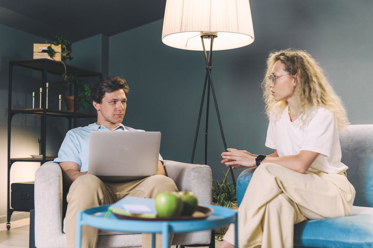 Two adults discuss work while sitting on sofas in a cozy, modern living room setting.